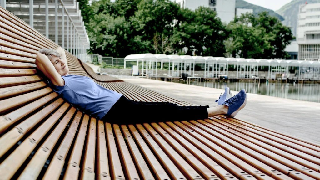 an older adult female in athleisure clothing rests on a curved bench outside with her eyes closed