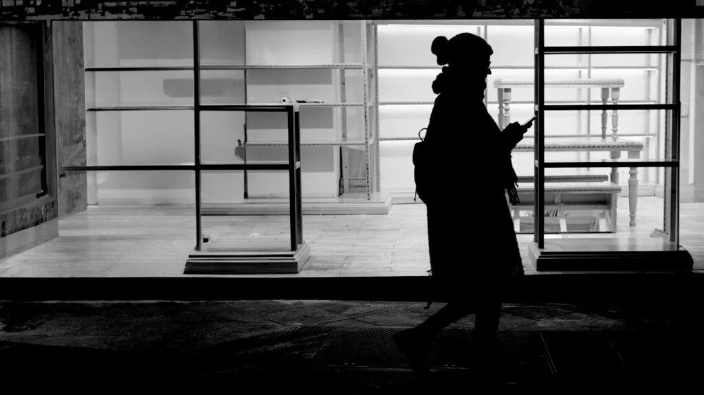 A silhouette of a woman wearing a hat and coat walking outside an empty store while looking at her phone at night